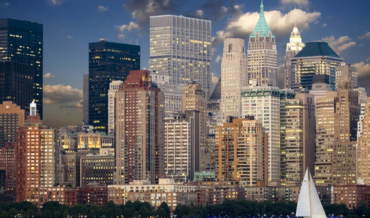 Panoramic view of New York City skyline with sailboat and river during twilight.